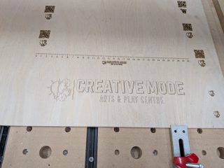 Plywood sheet on the CNC after engraving, showing the Creative Mode logo and an engraved centimetre scale